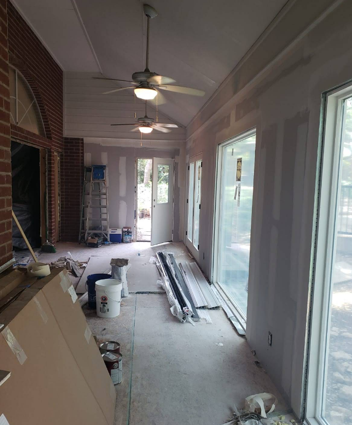 An interior view of a room under construction with exposed drywall, ceiling fans, a ladder, and large windows.