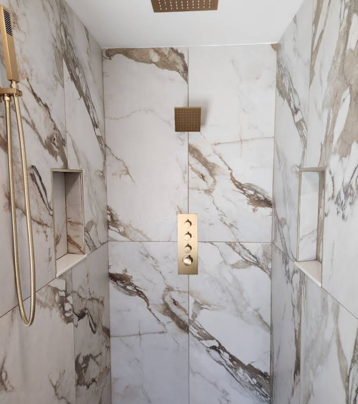 A shower stall with white and gold marble-patterned tiles, featuring gold fixtures, a rainfall head, and two niches.