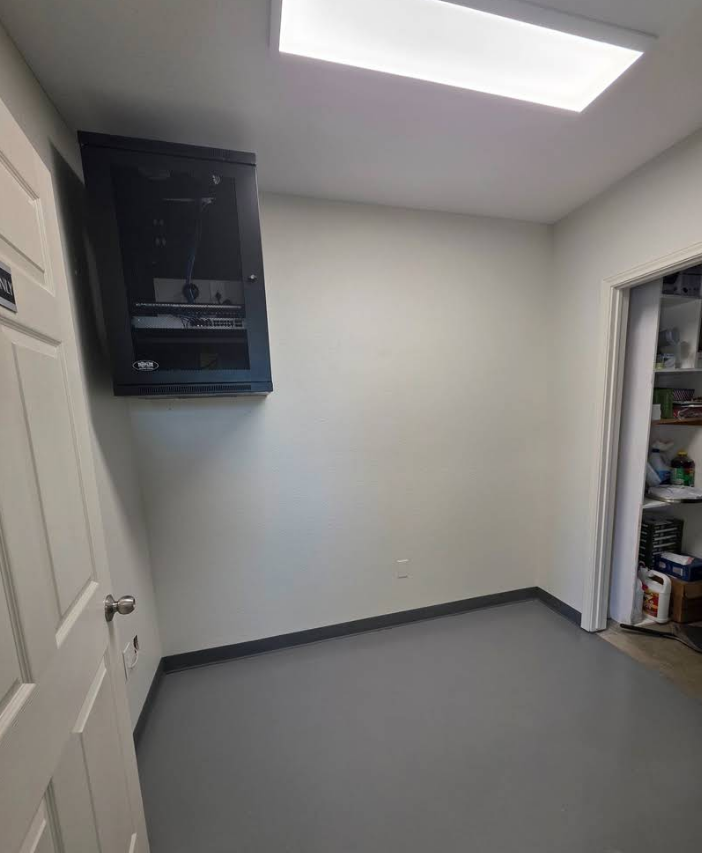 A small room with gray flooring, a black network cabinet mounted on the wall, and an open doorway to a cluttered closet.