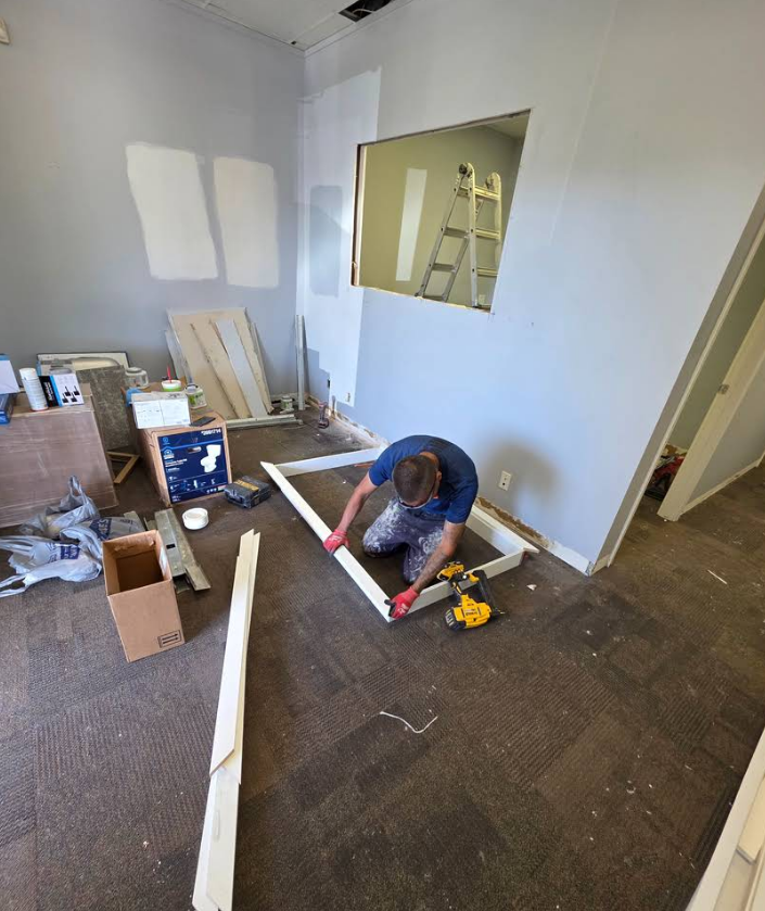 A person in red gloves kneels on a carpeted floor, assembling a rectangular wooden frame in a room under construction.