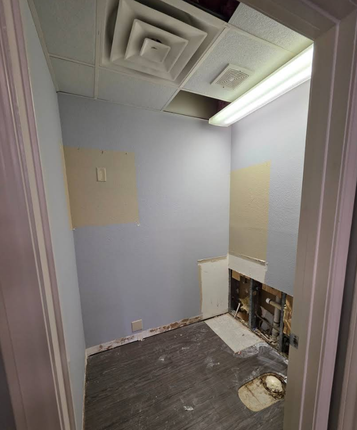 A small, unfinished room under construction with light blue walls, exposed plumbing, a ceiling vent, and gray flooring.