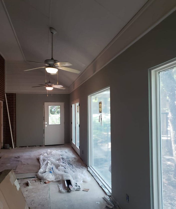An interior room under construction with grey walls, two ceiling fans, large windows, and floor protection plastic.