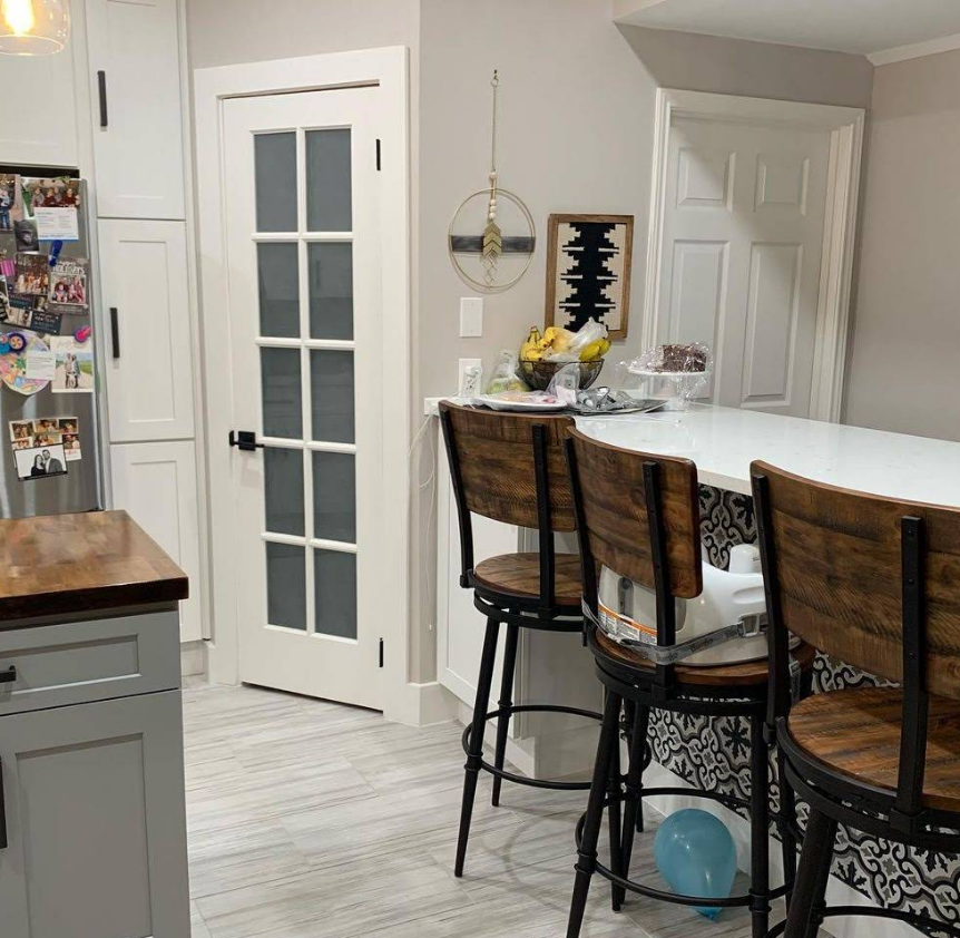 A kitchen with white cabinets, a glass-paneled pantry door, wooden bar stools at a counter, and gray tile flooring.
