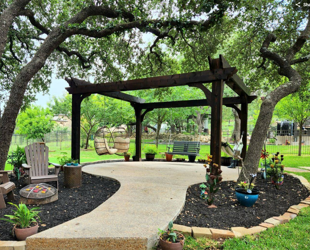 A wooden pergola stands on a stone patio in a lush backyard, featuring a hanging chair, garden bench, and potted plants.
