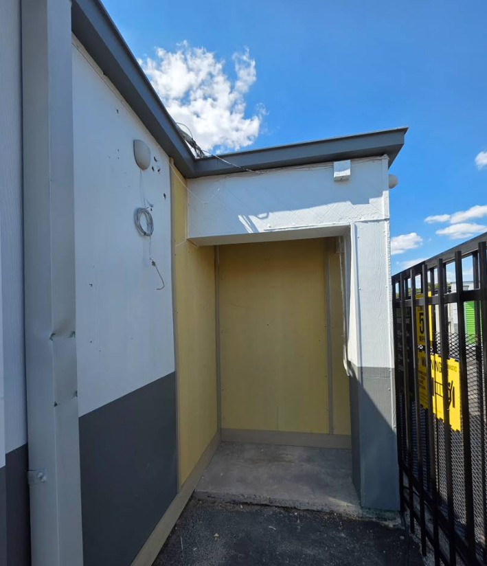 A sheltered, yellow-walled alcove next to a white and gray building, featuring an open entrance near a metal fence.