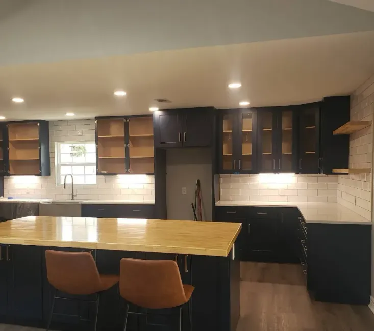 A modern kitchen features dark navy cabinets, white subway tile walls, a large wooden island, and two tan bar stools.