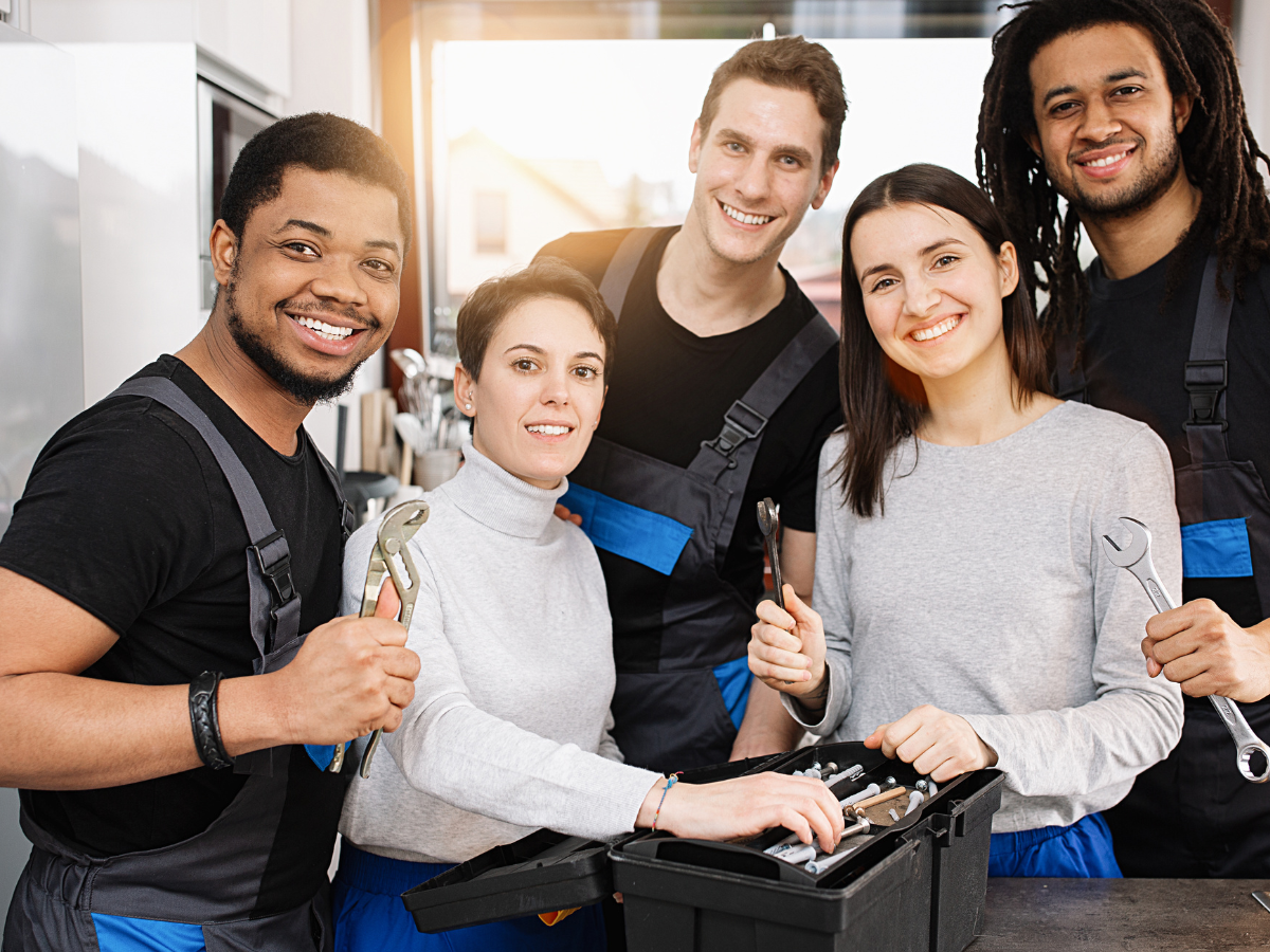 Group of diverse mechanics smiling, holding tools; garage setting.