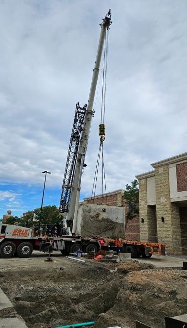 Crane lifting a large concrete block near a building under a cloudy sky.