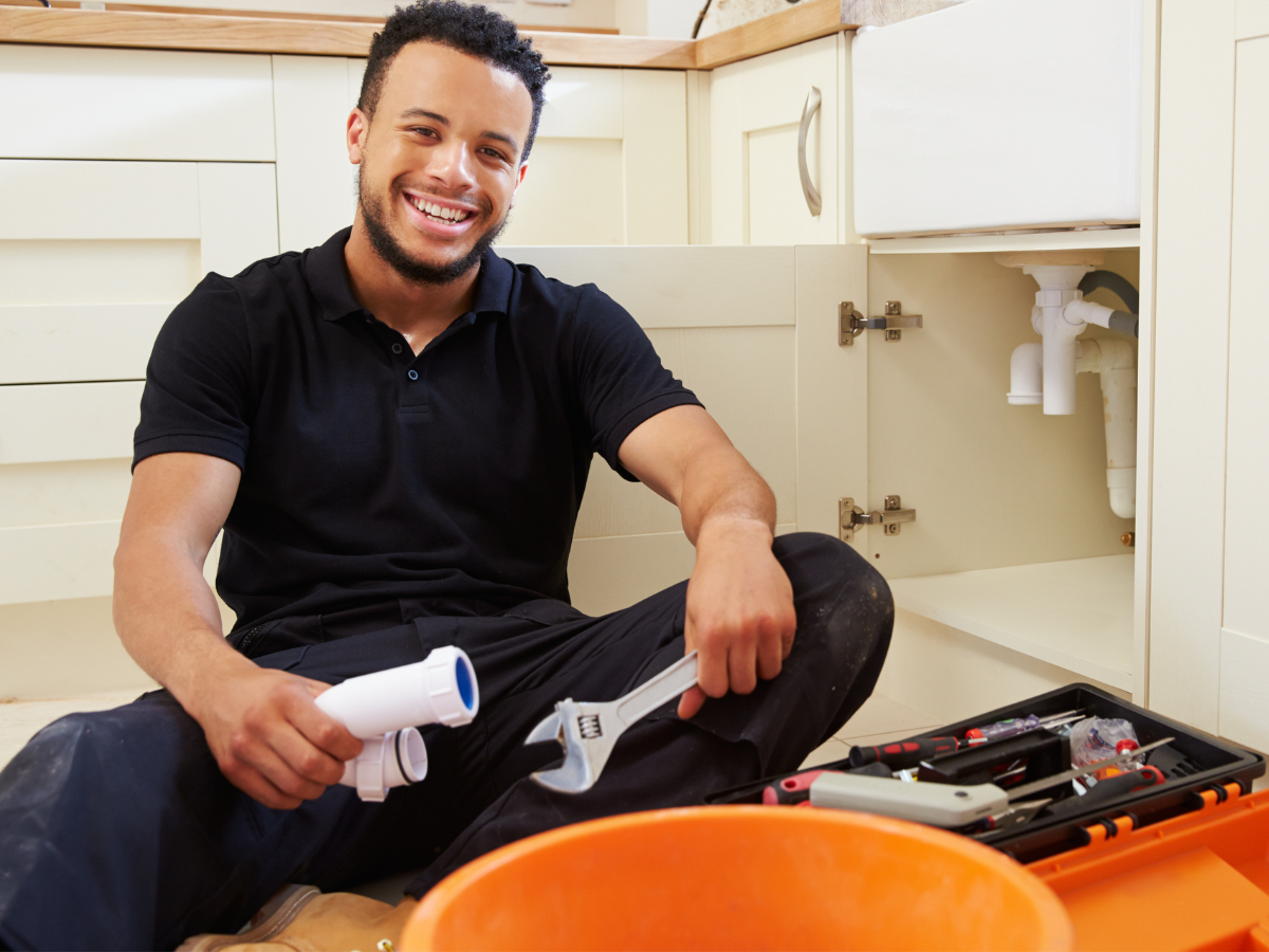 Man smiling, working on plumbing under a kitchen sink with tools and a bucket.