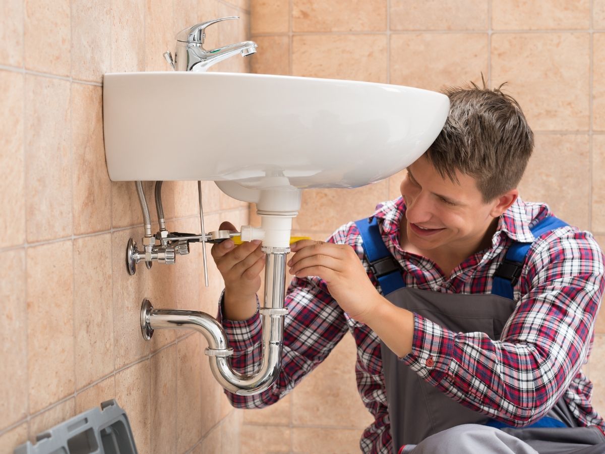 A plumber working under a kitchen sink, using a drain cleaning machine, with an open toolbox nearby.