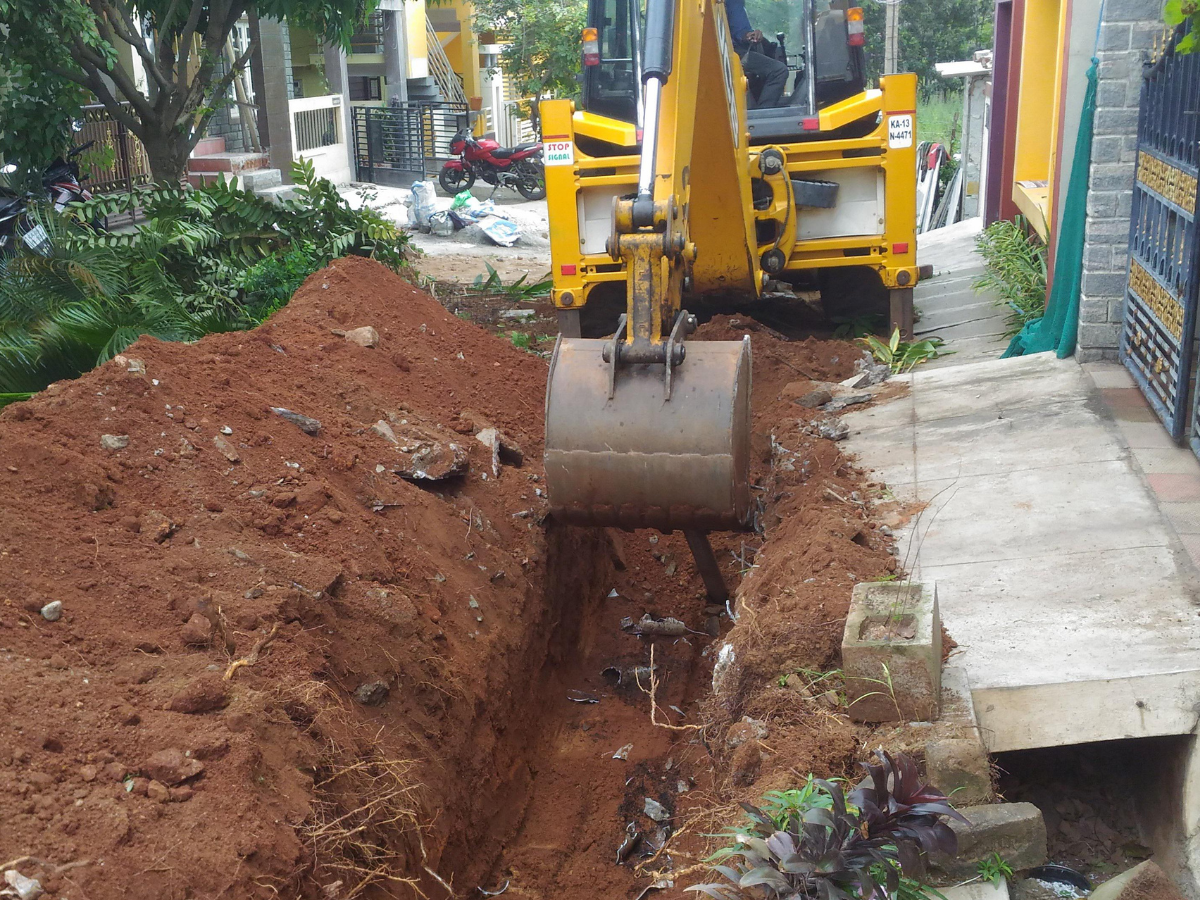Yellow excavator digging a trench next to a sidewalk. Red soil.