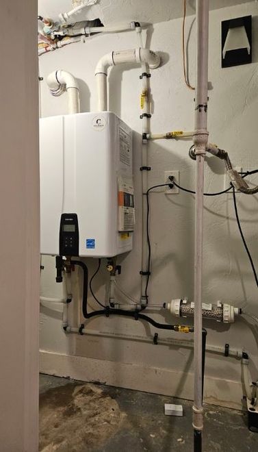 White tankless water heater on a basement wall with pipes, electrical, and a water filter.