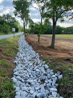 Rock-lined drainage ditch beside a road and grassy field with trees under a cloudy sky