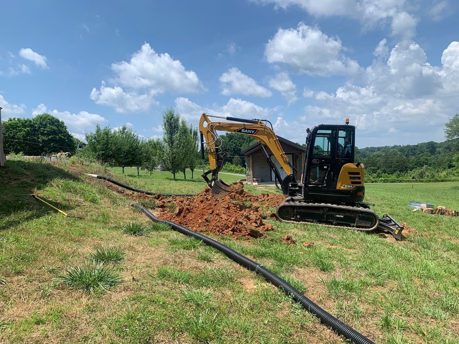 Excavator digging red soil near black pipes in a grassy field under a blue sky with clouds