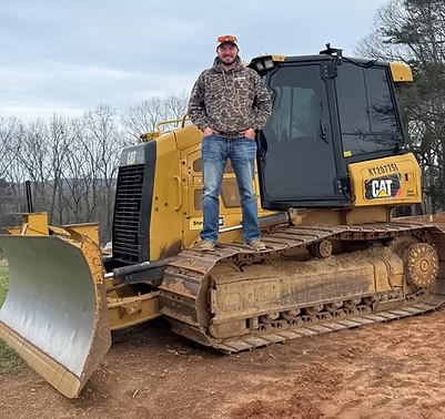 Man standing on a yellow CAT bulldozer on a dirt lot under an overcast sky