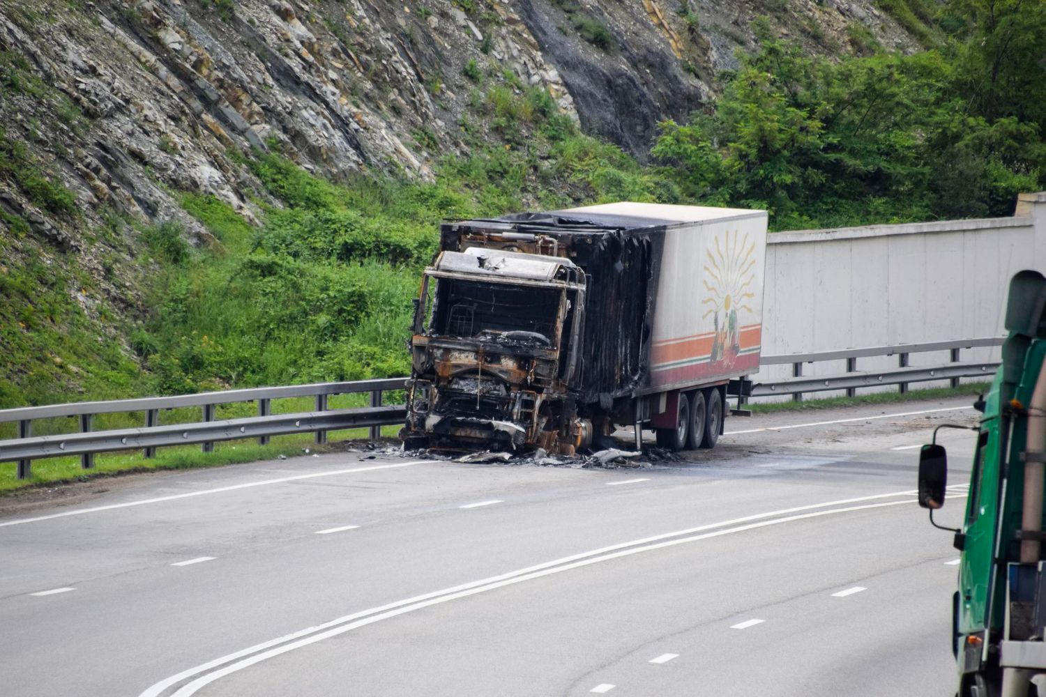 Burned semi-truck stopped on a highway shoulder beside a guardrail and rocky hillside.
