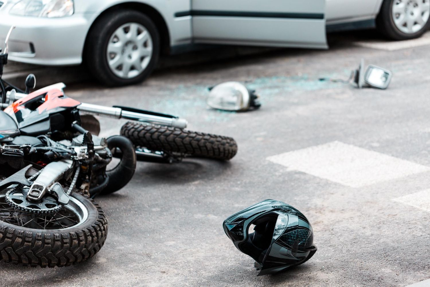 Motorcycle crash scene on a road with an overturned bike, scattered helmets, and a parked car nearby