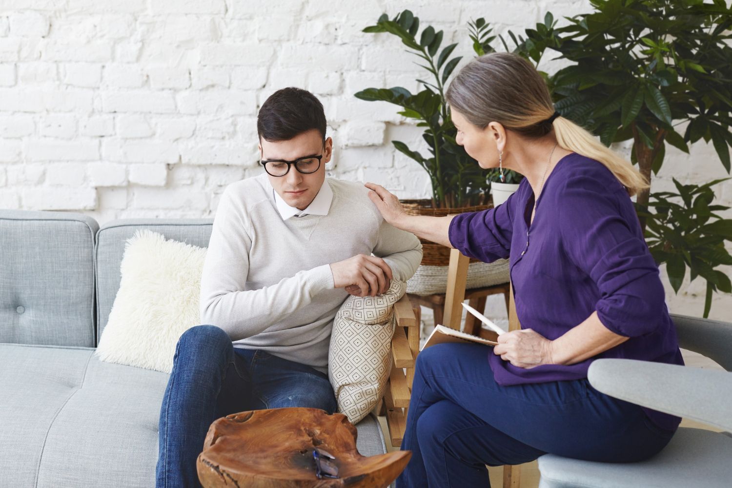 Two people on a couch pet a cat and dog in a bright living room