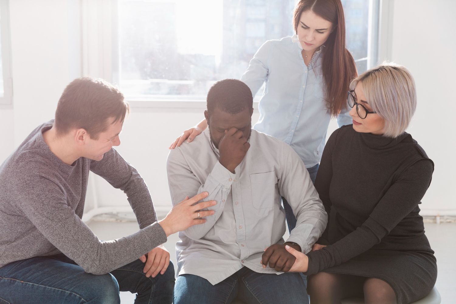 Four people sit together in a bright room, one comforting another with a hand on his shoulder.