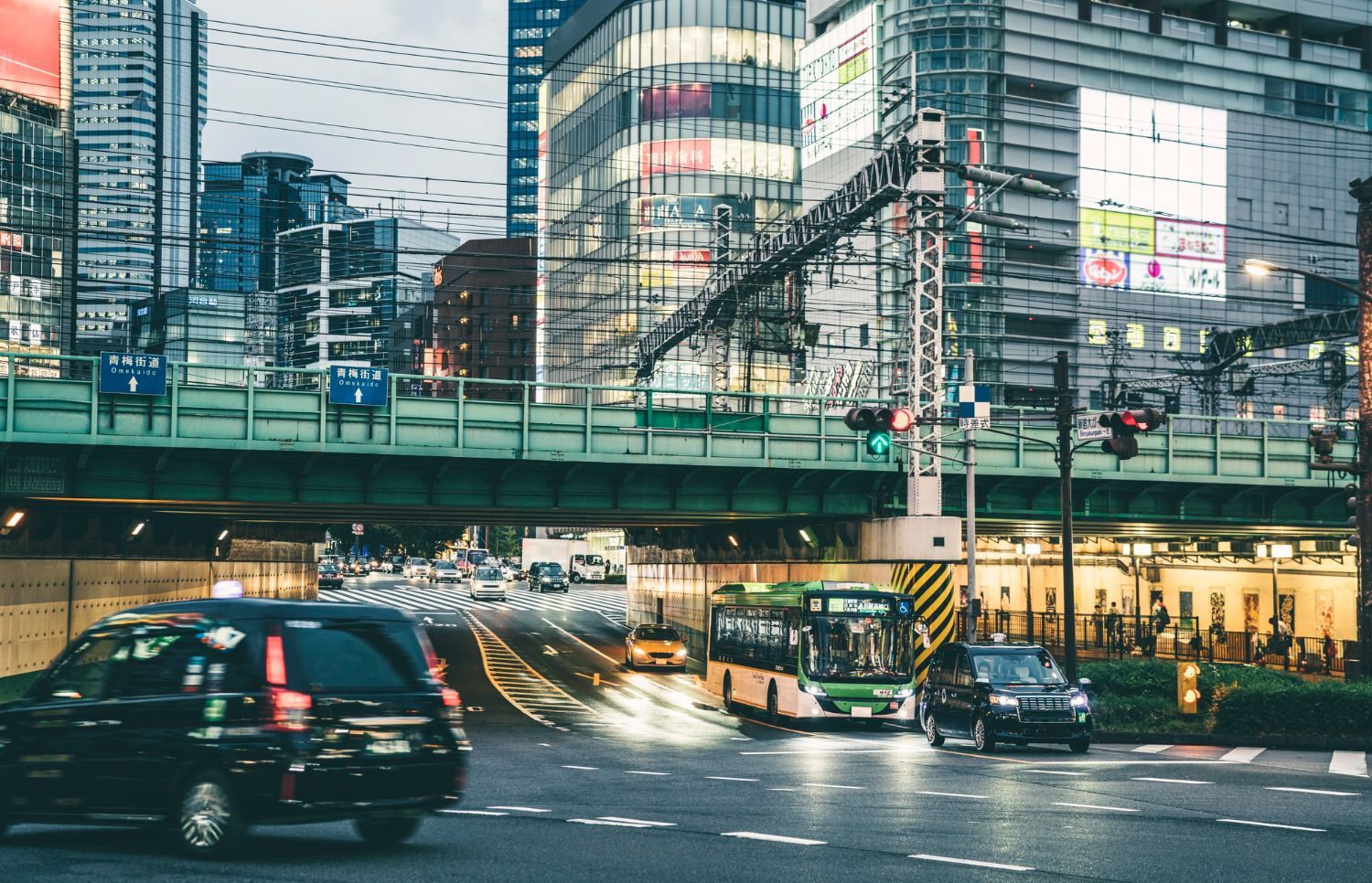 Busy city street at dusk with cars, buses, and illuminated buildings behind an elevated train track