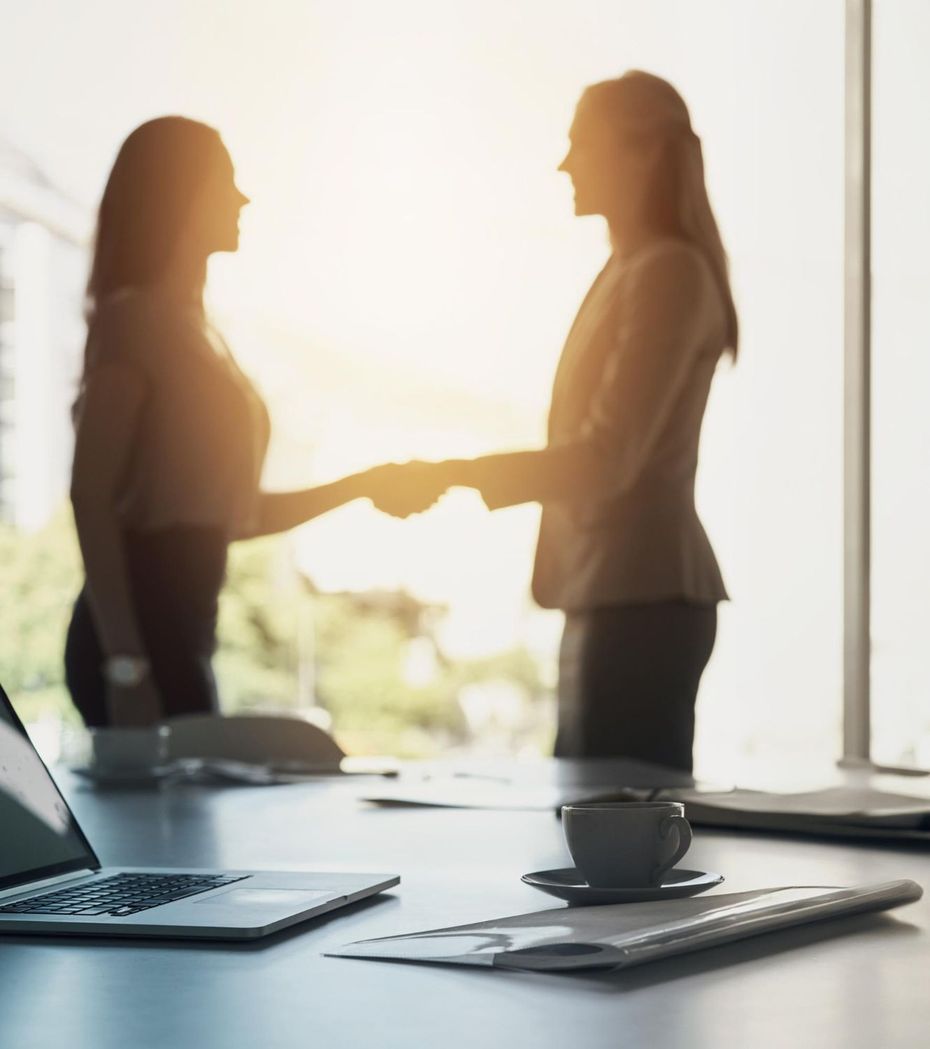 Two people shaking hands in a bright office, with a laptop and coffee cup on a desk.
