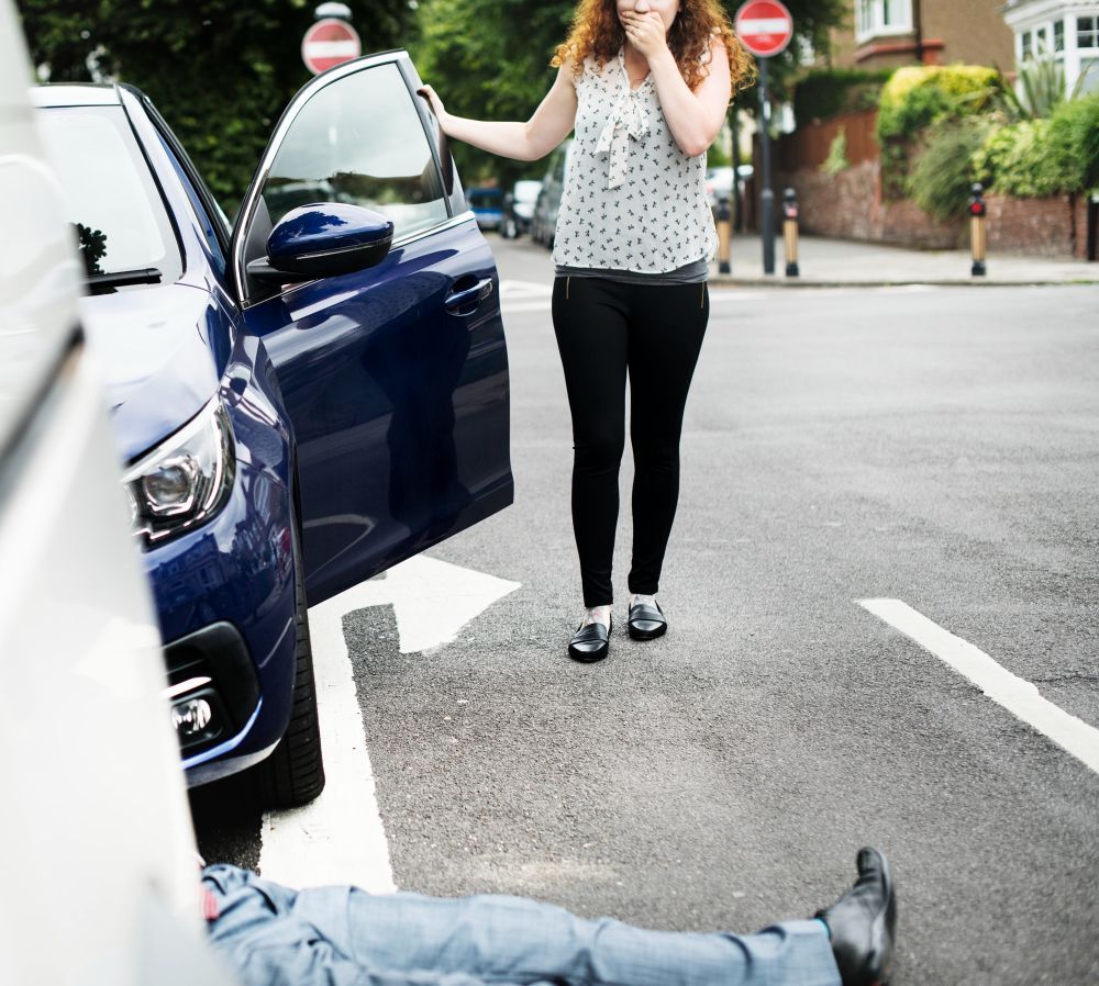 Person lying on street beside blue car while another stands nearby with hands over mouth.