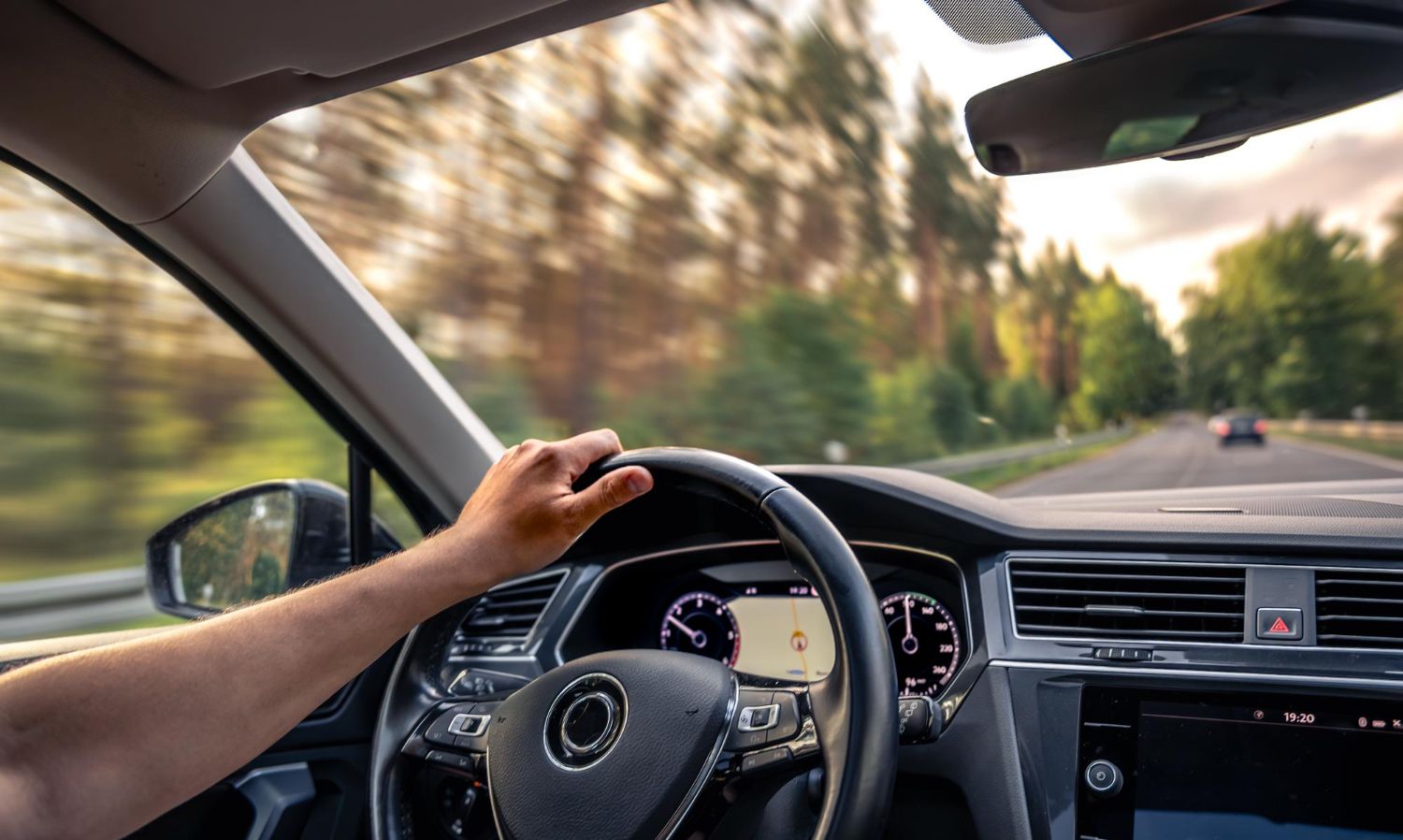 Driver’s hands on the wheel inside a car, driving past a blurred, tree-lined road at sunset