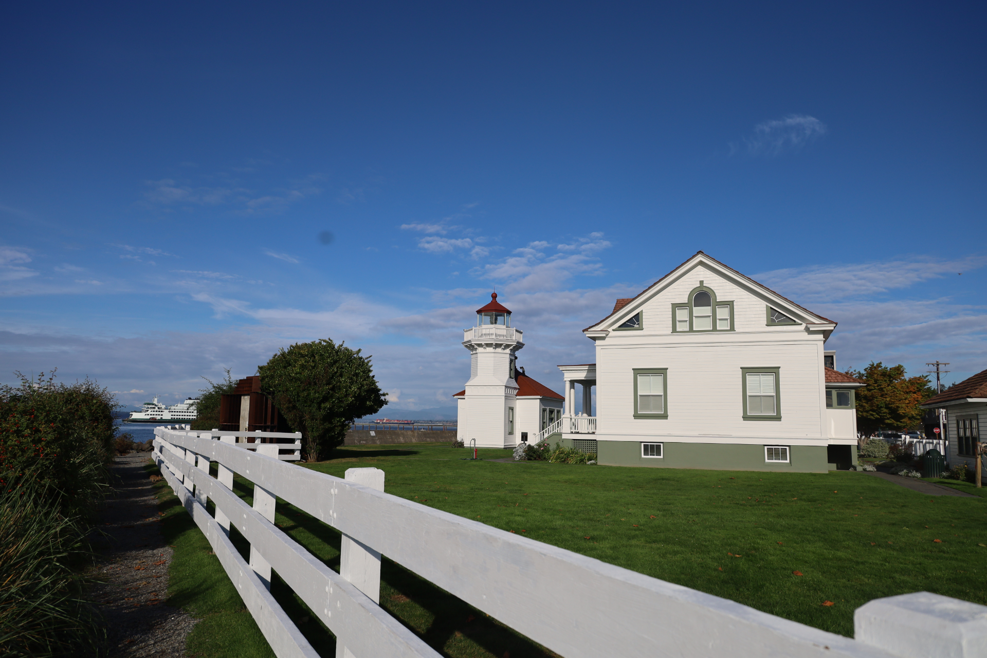 White lighthouse and house beside a white fence under a clear blue sky, overlooking the water
