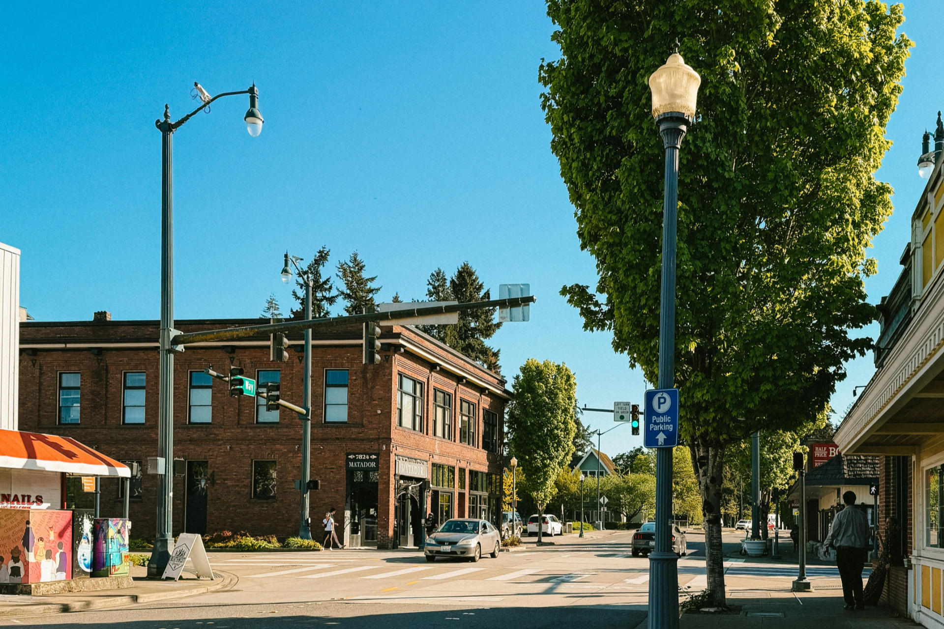 Sunny small-town street with brick buildings, trees, lamp posts, and a sidewalk café.