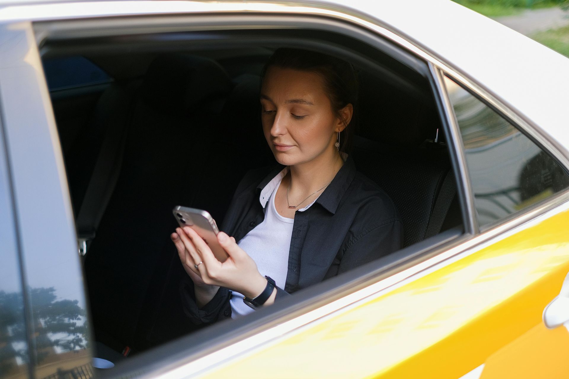 Person in a yellow car looks at a smartphone while seated by the window.