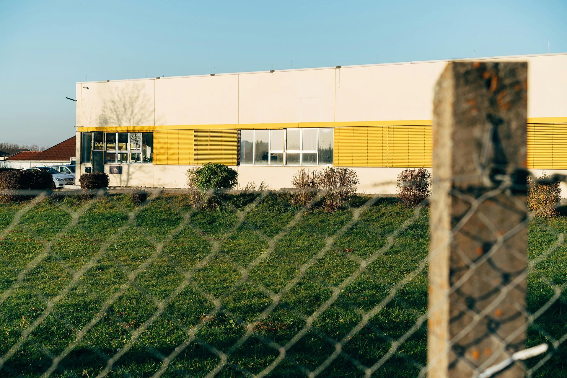 Industrial building behind a chain-link fence, with yellow siding and a grassy foreground.