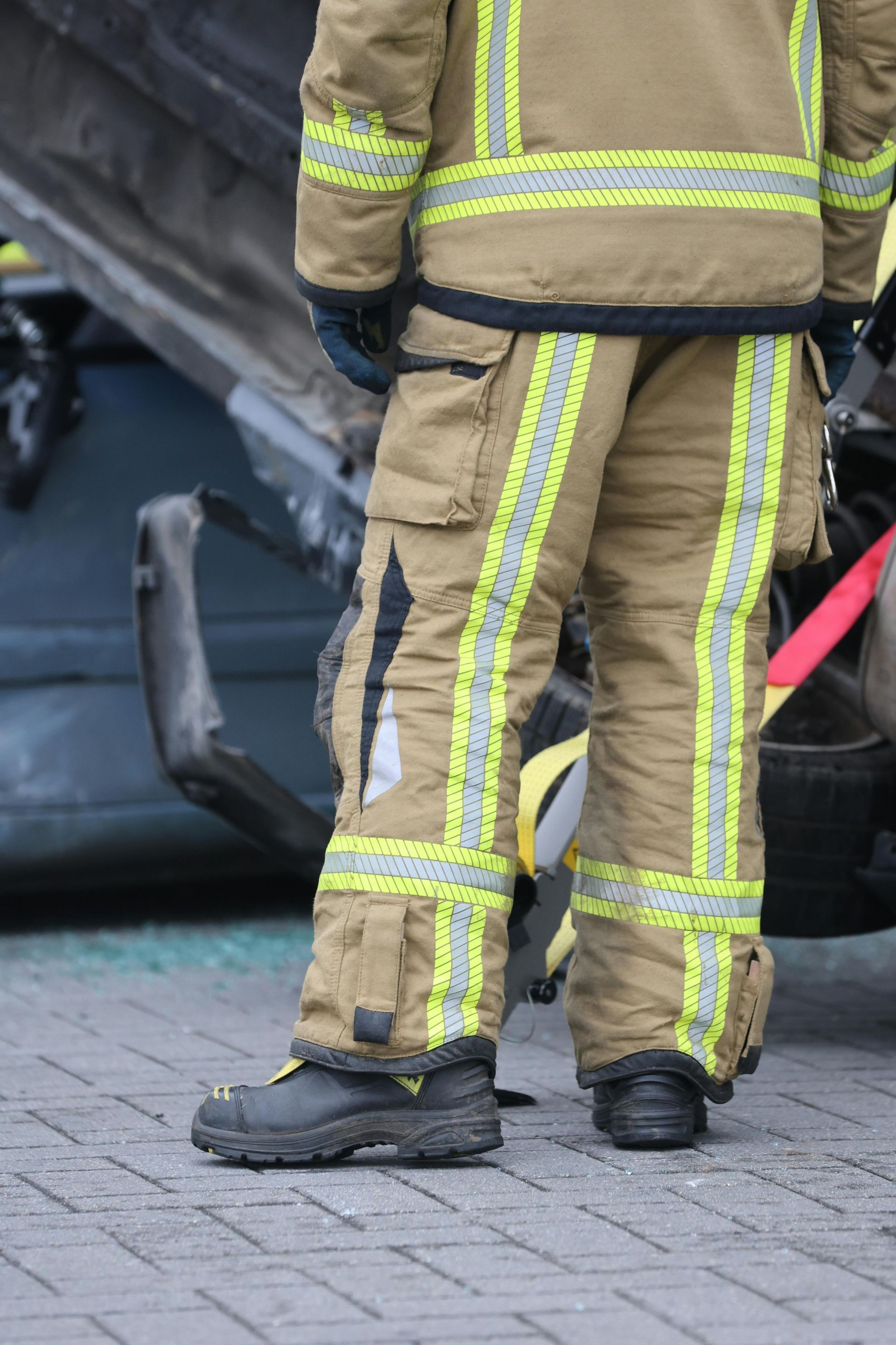 Firefighter in tan gear with reflective stripes standing beside a crashed vehicle on a paved street