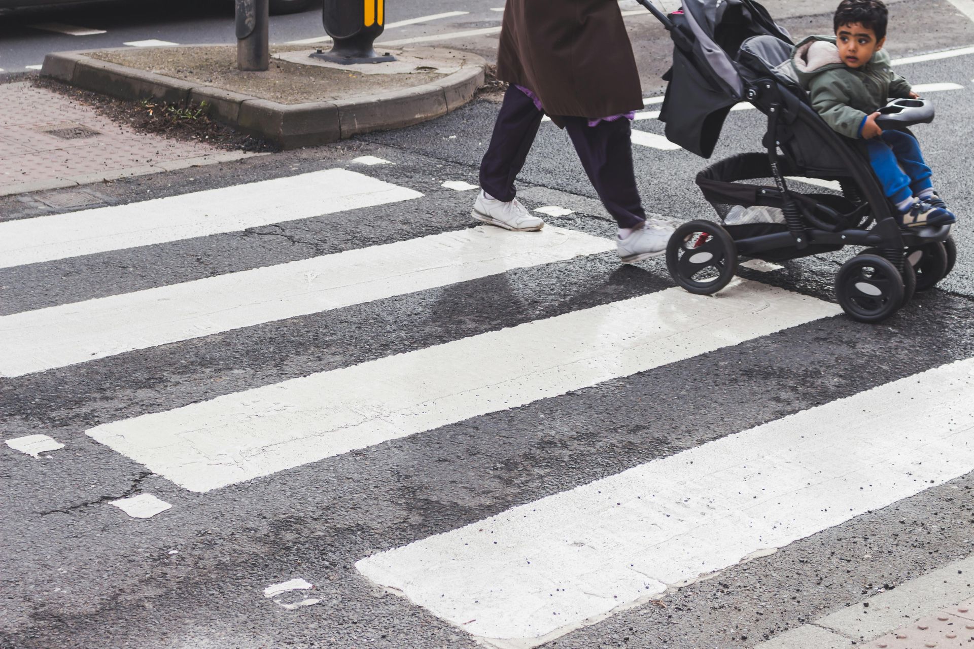 Person pushing a stroller across a crosswalk beside a child on a scooter