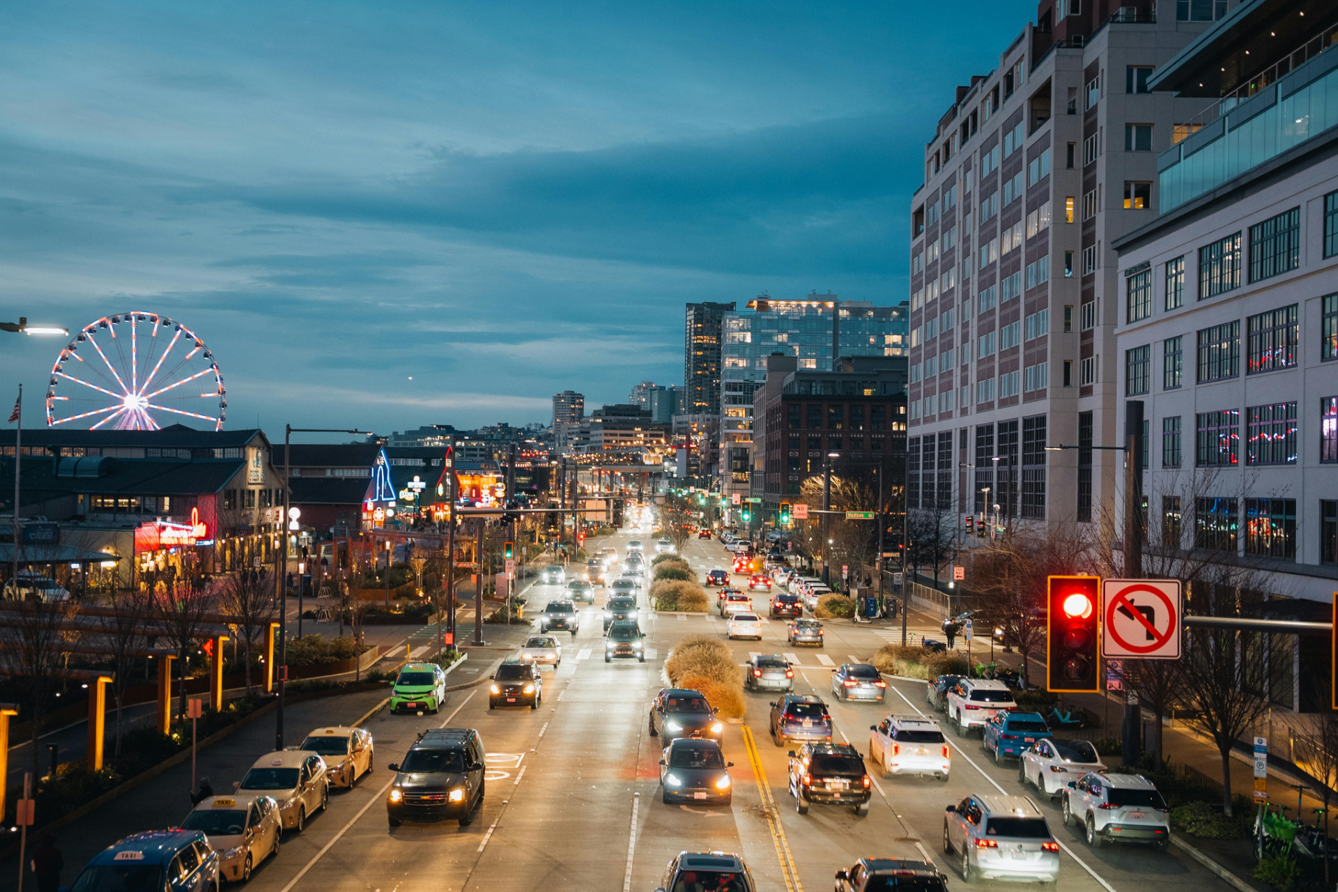 Busy city street at dusk with traffic, lit buildings, and a ferris wheel in the distance