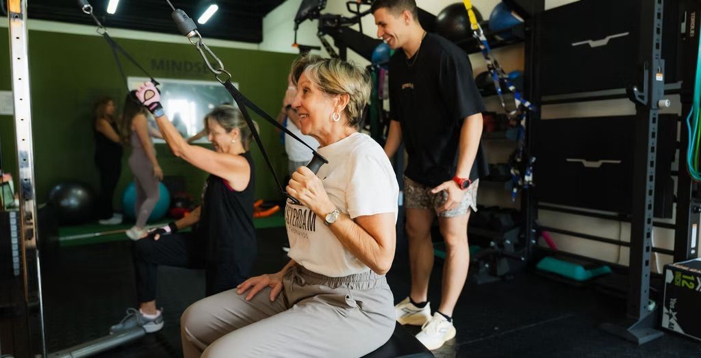 Varias personas haciendo ejercicio en un gimnasio; una de ellas levanta pesas mientras otras observan cerca.