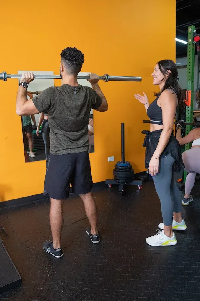 Dos personas en un gimnasio, una de ellas sosteniendo una barra con pesas sobre sus hombros junto a una pared naranja.