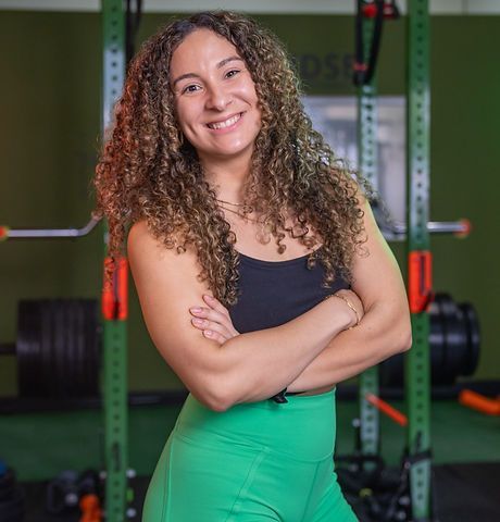 Mujer sonriente con cabello rizado de pie con los brazos cruzados en un gimnasio con pesas detrás de ella.