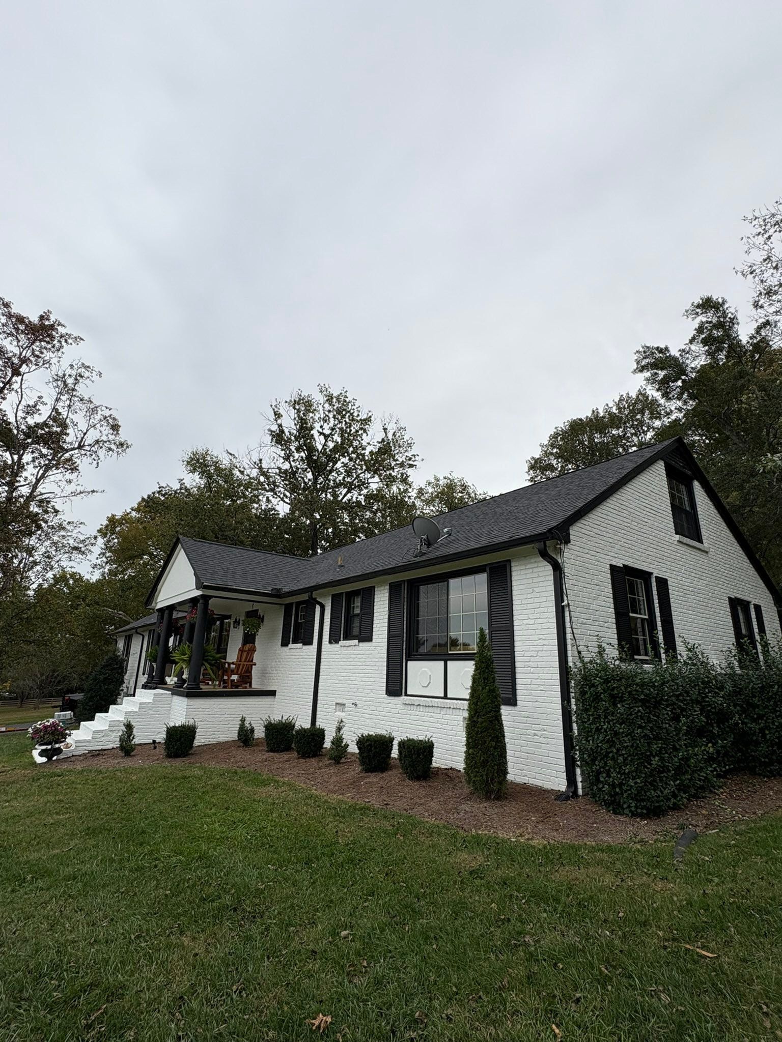 White cottage with dark roof on a grassy hill under an overcast sky, surrounded by trees and shrubs