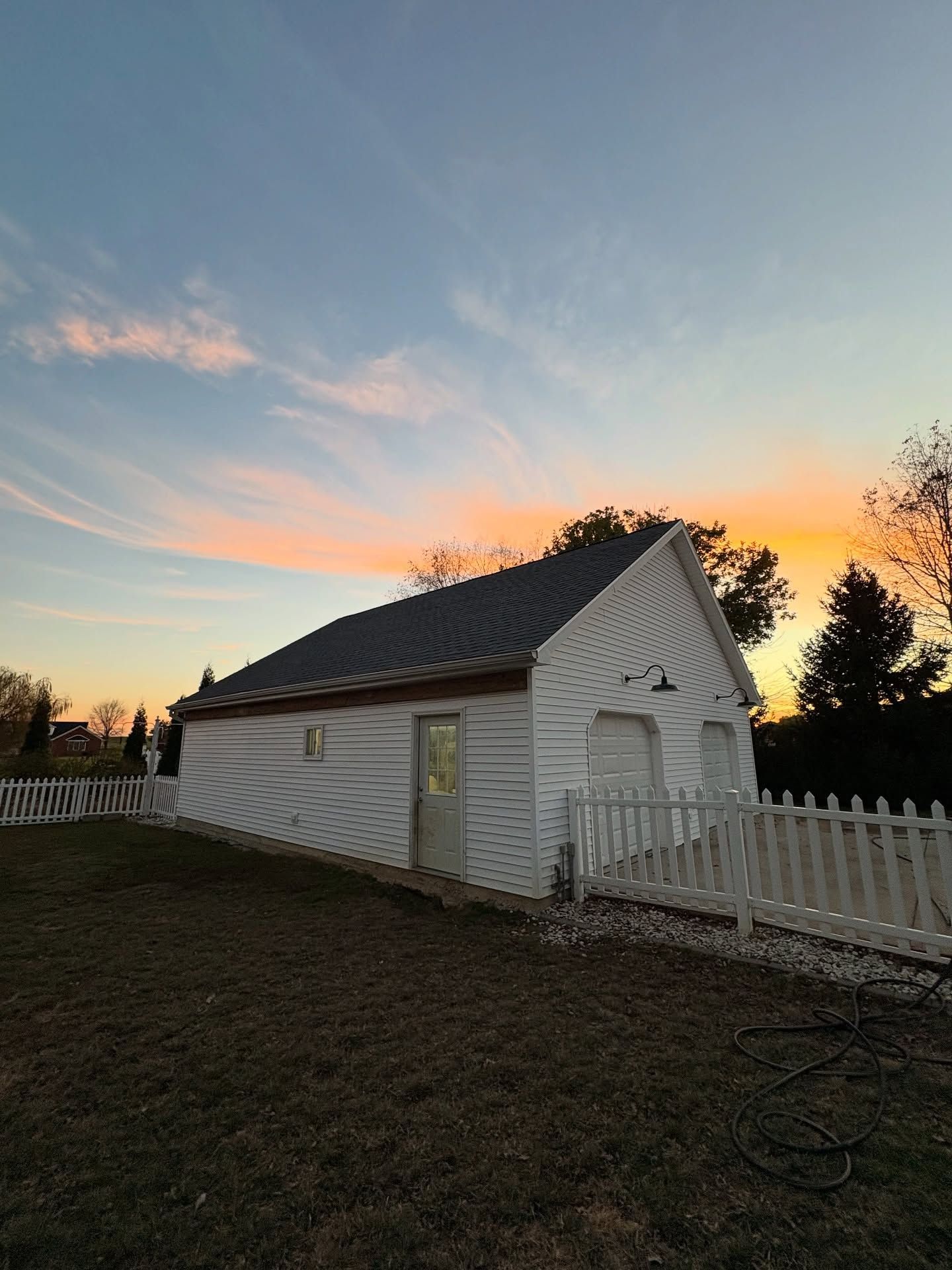 White barn-like shed by a picket fence at sunset, with a pastel sky and grassy yard.