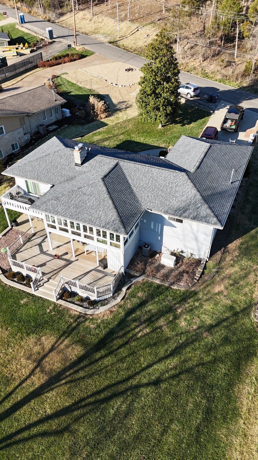 Aerial view of a white house with a gray roof, deck, and large yard beside a driveway and trees