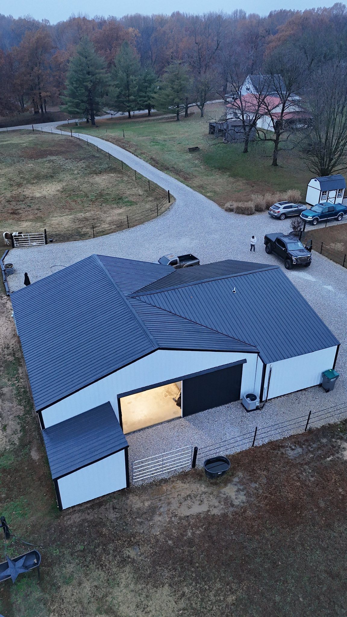 Aerial view of a gray-roofed house with a lit garage, gravel driveway, and wooded yard in autumn.