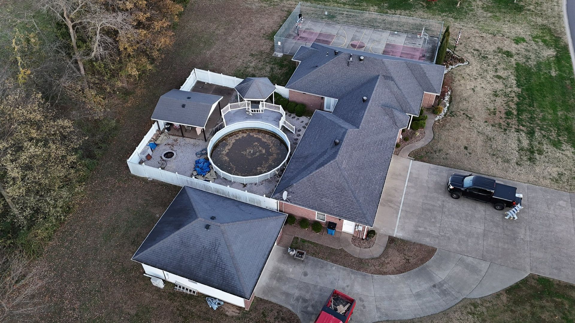 Aerial view of a house with a circular roof feature, driveway, and parked vehicles beside wooded land