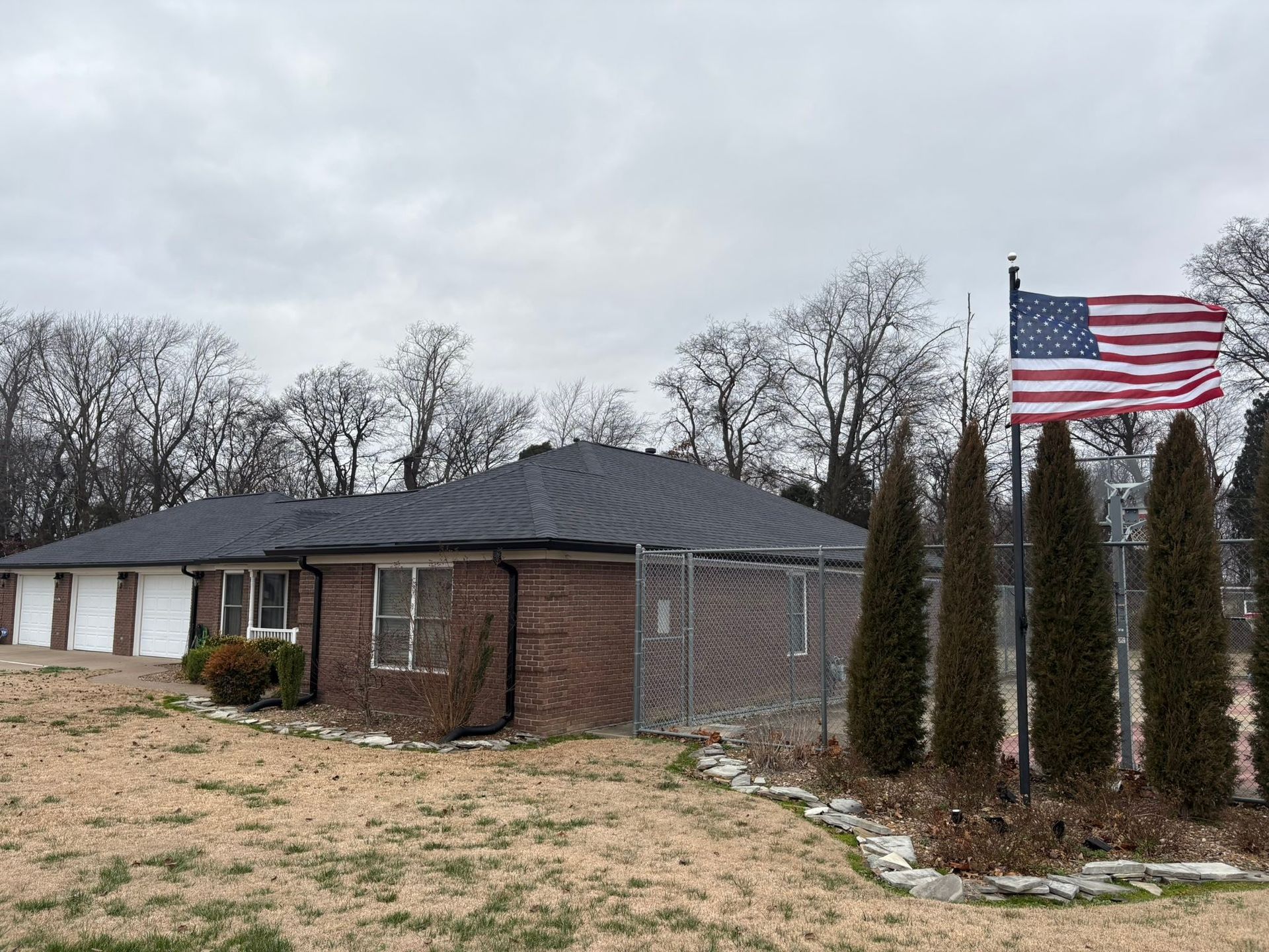 Single-story brick house with bare trees and an American flag in a cloudy yard