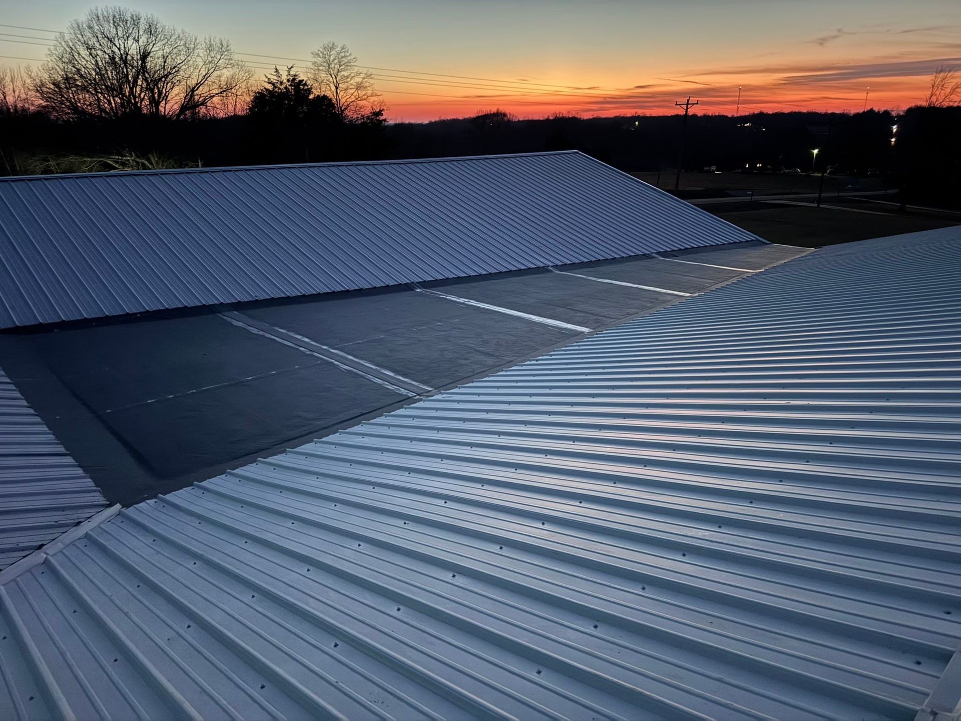 Sunset over corrugated metal rooftops with bare trees silhouetted against an orange-blue sky