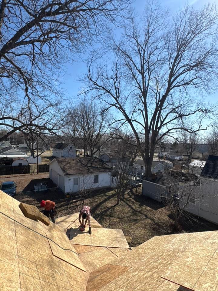 Roofers working on a tan shingle roof in a backyard with bare trees and neighboring houses.