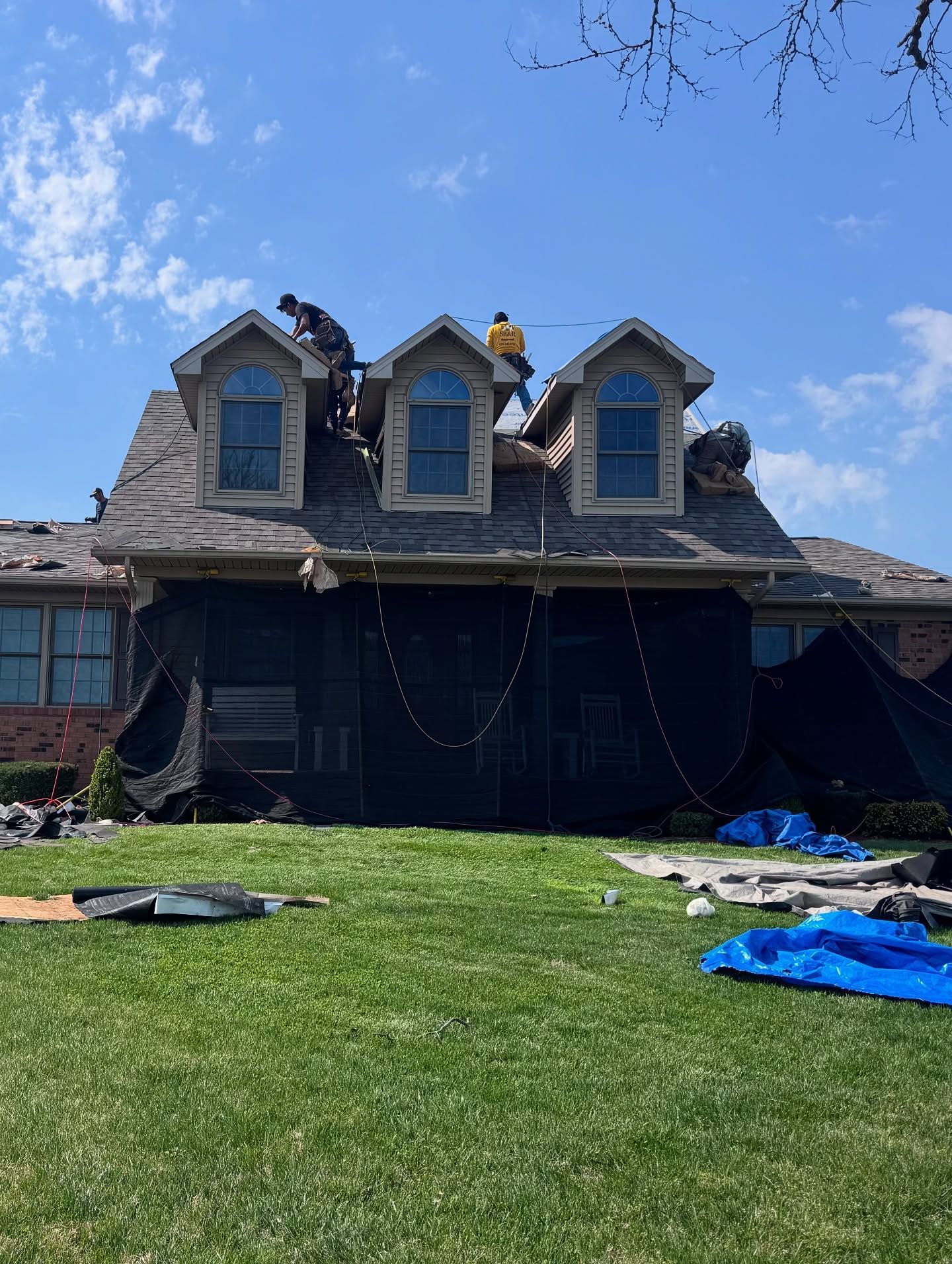 Roofers working on a house with dormer windows under a blue sky, with tarps on the lawn