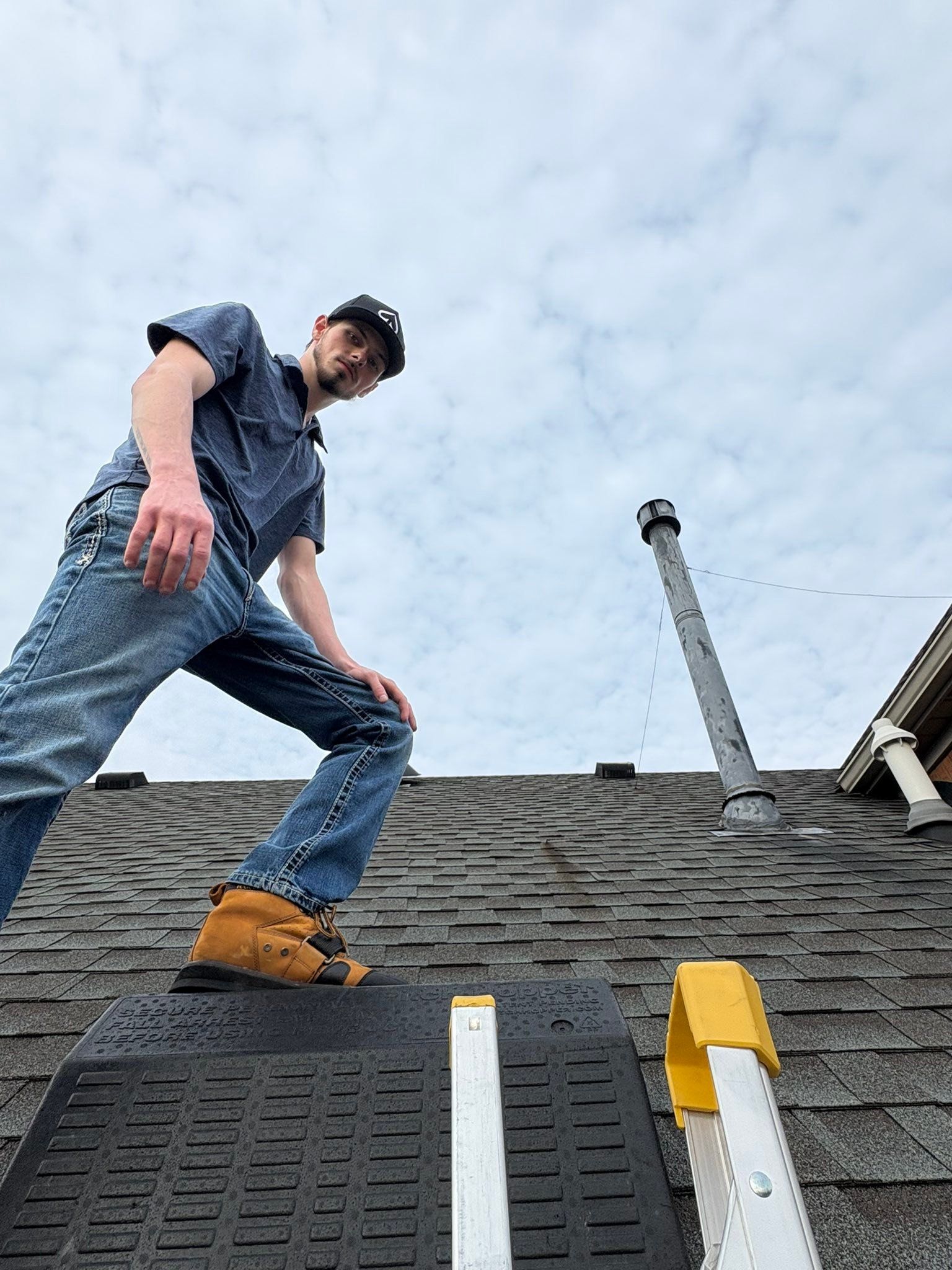 Man standing on a shingled roof under a cloudy sky, inspecting rooftop equipment with a white pipe and yellow vent.