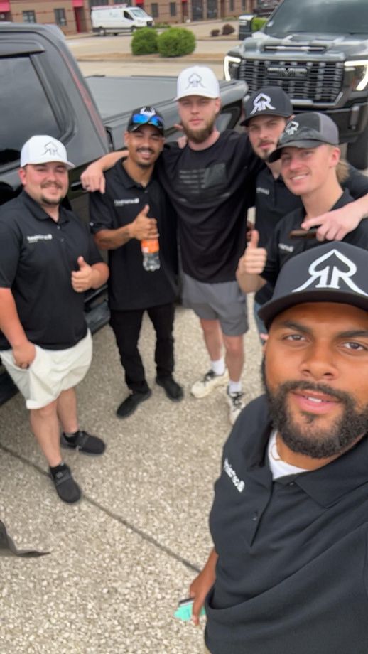 Five smiling friends pose outdoors in black shirts and caps, one taking the selfie in front.