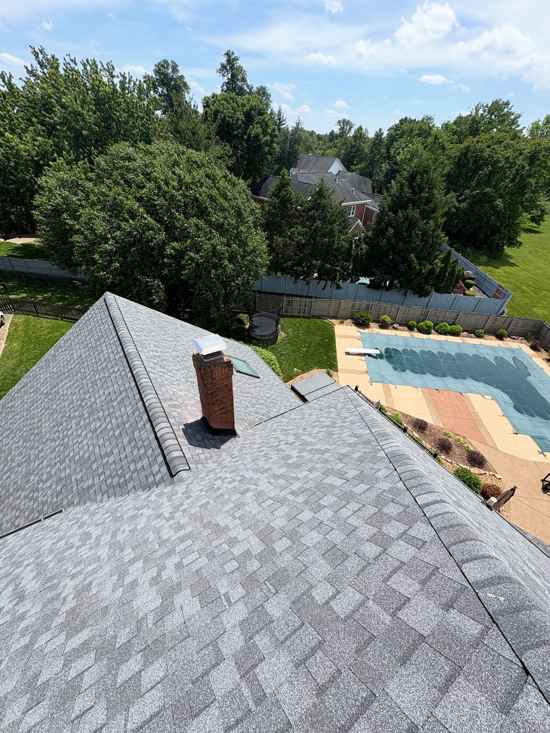 View from a shingled rooftop over a backyard pool, patio, and leafy trees on a sunny day