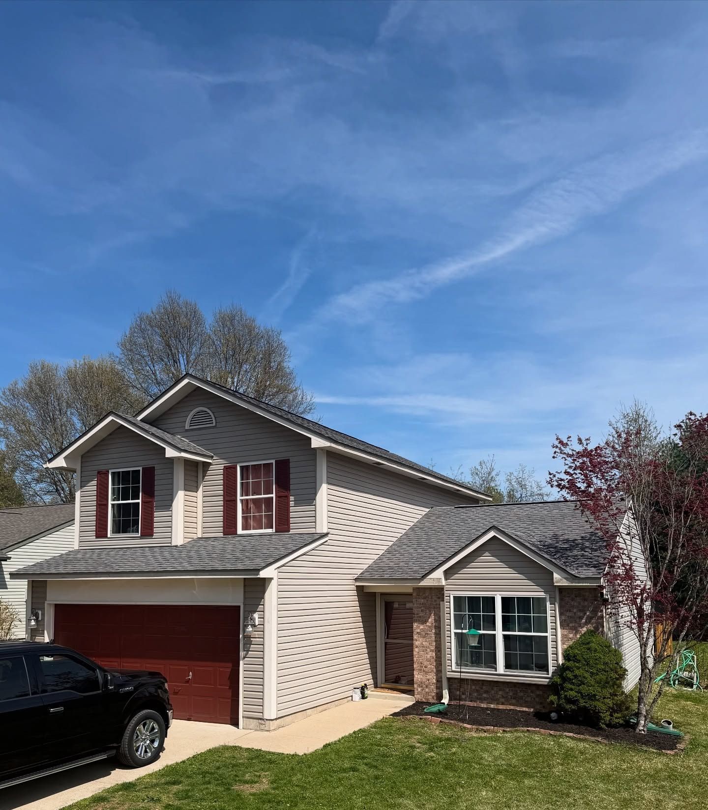 Suburban two-story house with beige siding, red shutters, a garage, and a black SUV in front under a blue sky.